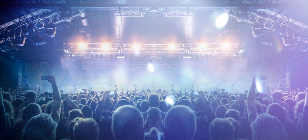 Image shot during a music festival. Light comes from a stage with a band show, people silhouettes are visible in front of it.