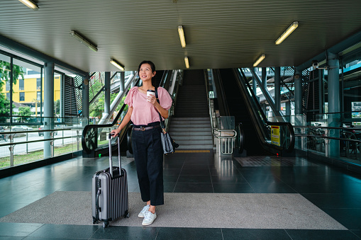 cheerful Asian woman with wheeled luggage standing at entrance of airport and having coffee, waiting for taxi.