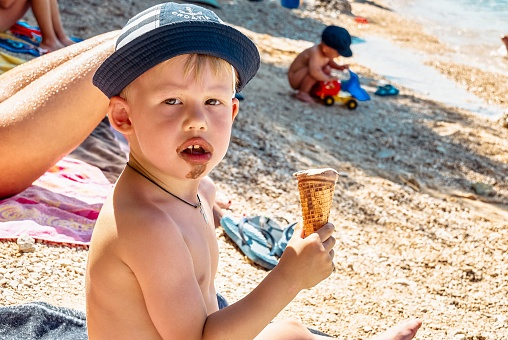 Cute little boy in panama hat eats chocolate ice cream sitting on beach. Toddler with dirty mouth enjoys summer vacation in Omis close view