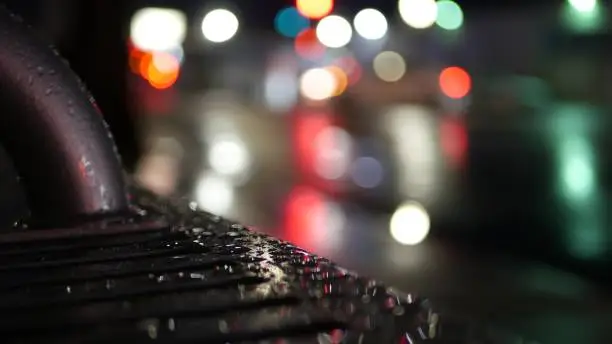 Photo of Car traffic lights reflection on bus stop bench. Water rain drops on wet metal.