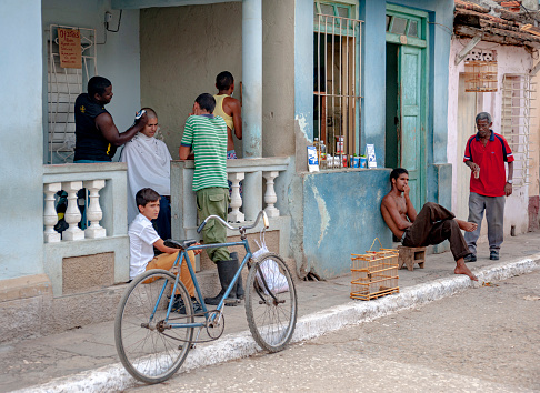 A lot of things happening in a barbershop on the front porch of a house and next to a grocery store of food items in Trinidad, Cuba.