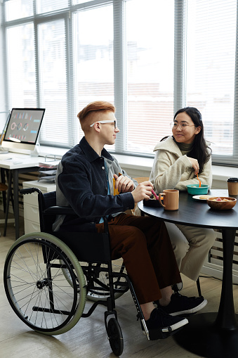 Vertical full length portrait of creative young man in wheelchair enjoying lunch in office with coworker