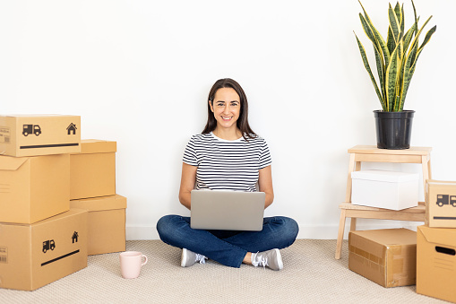 Happy young woman smiling at camera while using laptop computer in new home - Moving and relocation concept