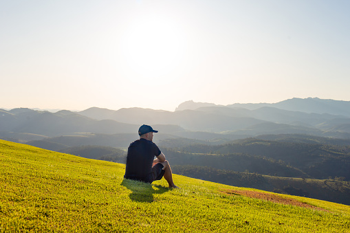 Young man sitting on the grass and watching the sunset in the mountains in the background.