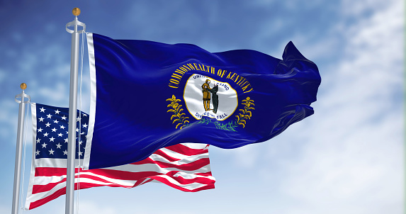 The Kentucky state flag waving along with the national flag of the United States of America. In the background there is a clear sky. Kentucky is a state in the Southeastern region of the United States