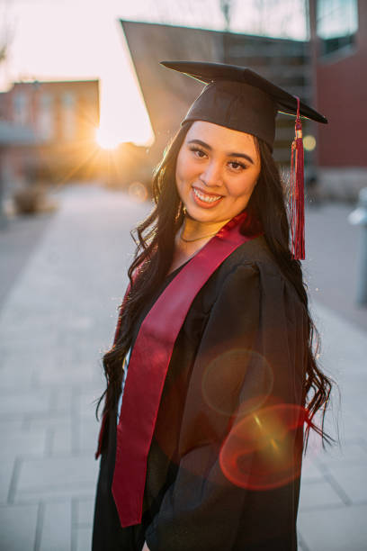 Portrait of a Cheerful Young Hispanic Woman Wearing a Cap and Gown Looking Over Her Shoulder with a Maroon Sash in the Spring Before Graduation stock photo