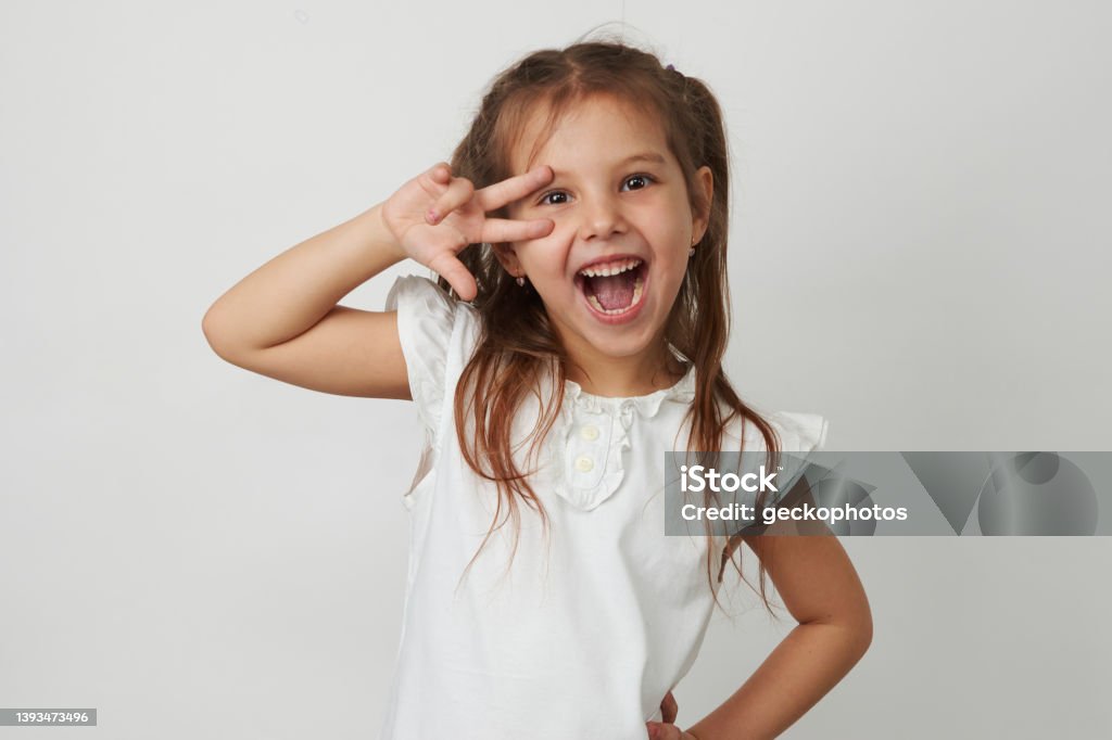 Portrait of cute girl give v-sign near eye Portrait of cute girl give v-sign near eye. Smiling little girl posing over white background with copy space, close-up Child Stock Photo