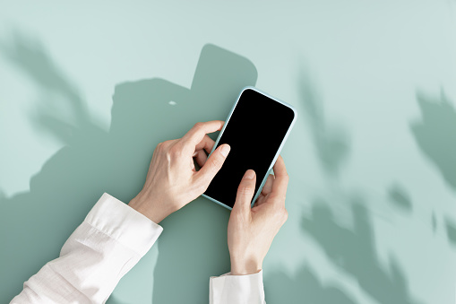 Woman using phone with black blank screen, minimal flat lay on mint blue background. Smartphone mock up. Digital online summer lifestyle composition with leaves shadows and sunlight, pastel colors