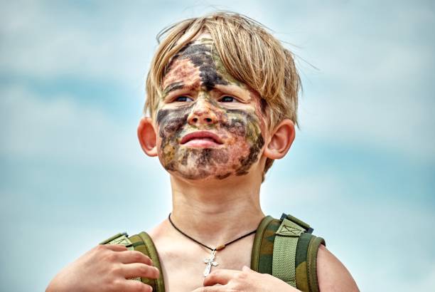 Toddler with dirty painted face and camouflage backpack stands on beach against sky. Cute blond boy tourist explores nature