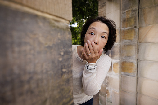 Woman with a scared expression looking around the corner of a stone wall.