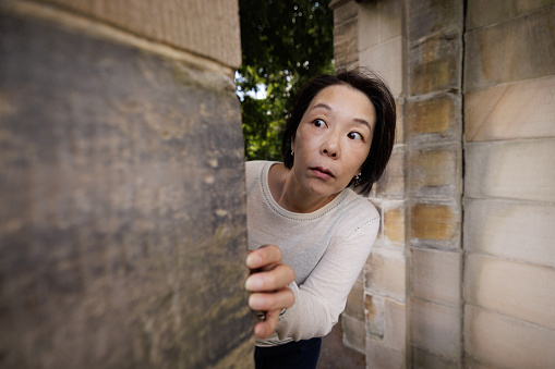 Woman with a scared expression looking around the corner of a stone wall.
