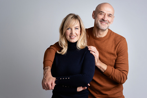 Studio shot of a happy middle aged couple standing together at isolated background with copy space. Happy mand embraching beautiful blond haired woman.