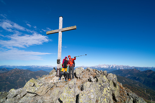 Mountaineer couple standing together close to summit cross on mountain peak on sunny but cold and windy day in autumn, man pointing with hiking pole, mountain range panorama in background