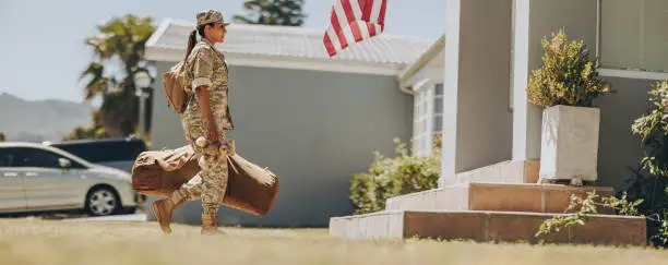 Courageous military mom holding a teddybear while walking towards her house with her luggage. American female soldier coming back home after serving her country in the army.