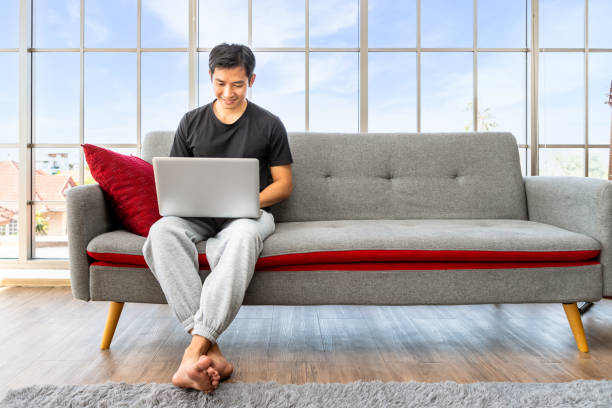istock Happy young Asian man using laptop computer sitting o sofa in living room, work from home concept. 1393558035