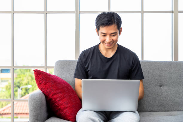 istock Happy young Asian man using laptop computer sitting o sofa in living room, work from home concept. 1393558055