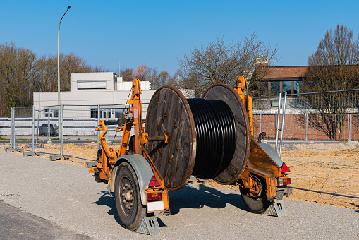 A wooden reel with an electric cable is mounted on a two-wheeled trailer. Construction site. Buildings in the background.