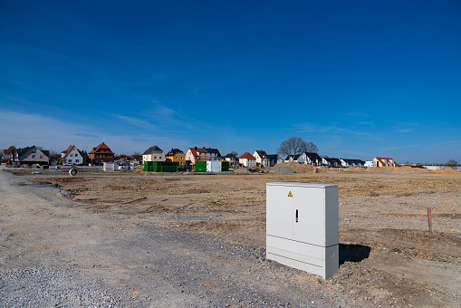 Distribution electric box of white color at the construction site. In the background are residential buildings and construction equipment.