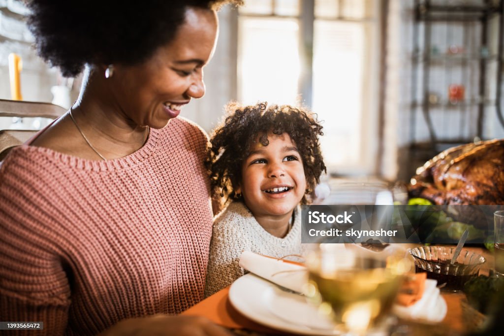 Happy black single mother and her daughter on Thanksgiving meal at dining table. Happy African American mother enjoying with her daughter during Thanksgiving meal at dining table. Thanksgiving - Holiday Stock Photo