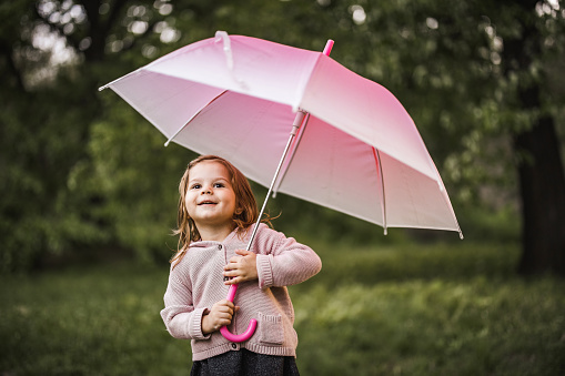 Small girl standing in the park with pink umbrella and looking away.