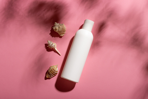 White cosmetic bottle with face cream or lotion and telana against a pink background with seashells and shadows. Sun cream, summer cosmetics.