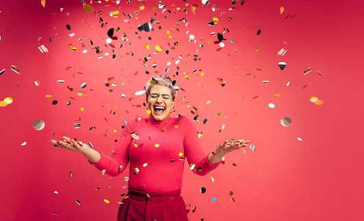 Celebrating with confetti. Carefree young woman getting excited while standing under falling confetti in a studio. Vibrant young woman having fun and celebrating life against a red background.