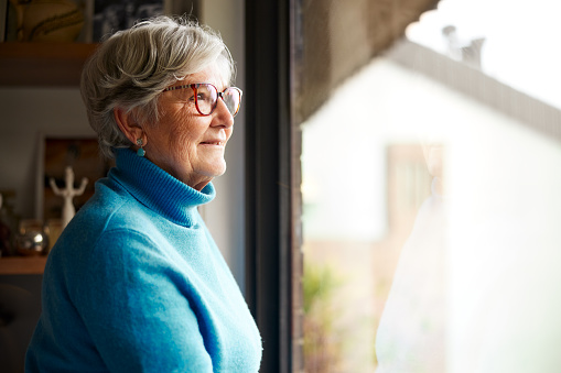 An elderly woman stands next to a window and looks outside.\nView with copy space.