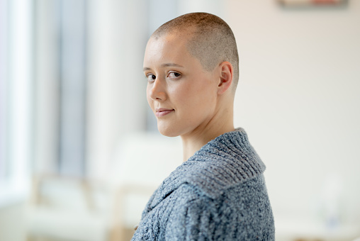 A young woman in her 20's sits on a sofa as she poses for a casual portrait.  She has her head freshly shaven, is wearing a comfortable sweater and is looking over her shoulder with a neutral expression.