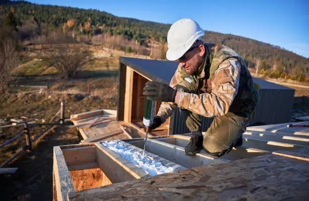 Photo of Male builder doing thermal insulation on roof of wooden frame house by polyurethane foam.