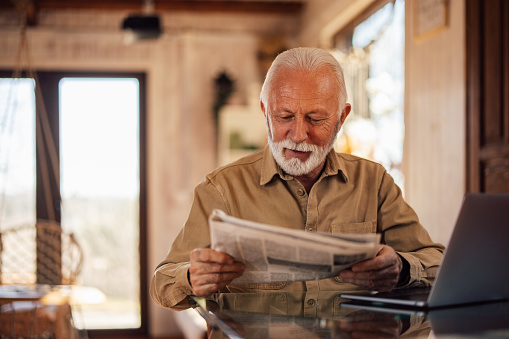 Mature man enjoying alone at home, holding and reading a newspaper.