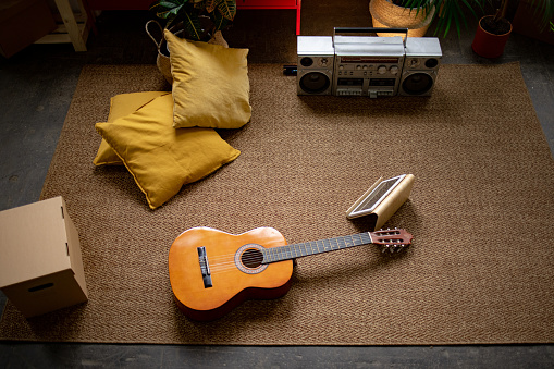 High angle view of an acoustic guitar, tablet computer, boom box, yellow pillows, and other common house items sitting on the apartment floor on the carpet, no people