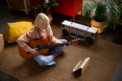 High angle view of a young woman with blonde hair playing an acoustic guitar at home while sitting on the floor of her loft apartment and using a tablet computer to watch guitar tutorials on the internet