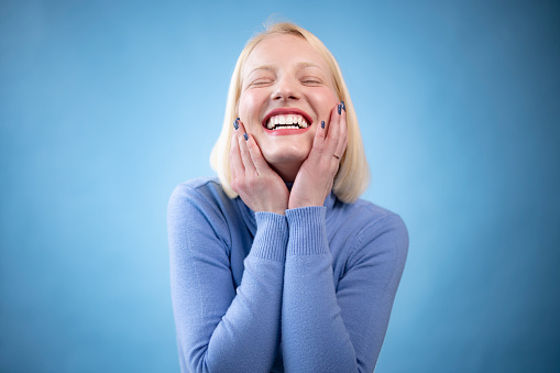 Portrait of a cheerful young blond hair woman wearing a blue turtleneck shirt standing in a studio with a blue background radiating joy around her with a big toothy smile and holding her own cheeks with her hands while having her eyes closed