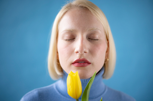 Close up of a young blond hair woman wearing a blue turtleneck shirt standing in a studio with a blue background holding a yellow tulip flower and having her eyes closed
