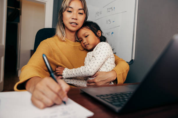 joven madre trabajadora escribiendo notas mientras abraza a su hija - trabajando en casa fotografías e imágenes de stock