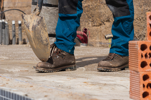 Worker's legs in construction site with a shovel on safety boots and bricks