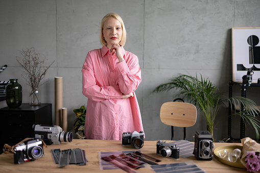 Portrait of a young female photographer standing in her studio apartment, having her camera equipment on a table in front of her and looking at camera