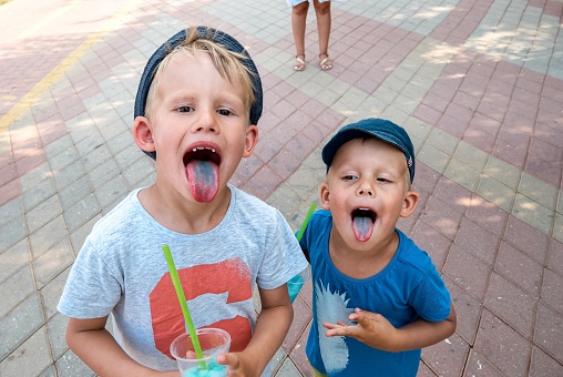 Preschooler and toddler brothers with blue tongues from eating fruit ice. Cute boys in hats stand on street in shadow enjoying summer vacation closeup