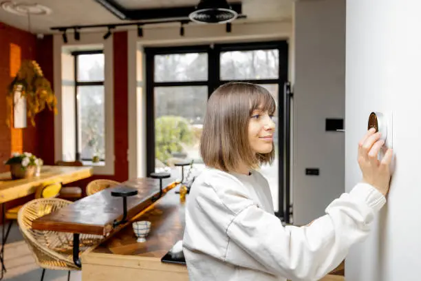 Woman controlling home temperature with electronic thermostat mounted on the wall in living room at home. Concept of modern technologies and smart home