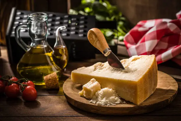Front view of a crumble and grated parmigiano reggiano surrounded by a grater, cherry tomatoes, olive oil and basil on a rustic wooden table.