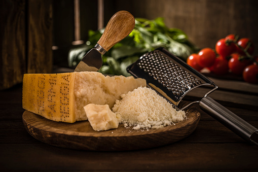 Front view of a crumble and grated parmigiano reggiano surrounded by a grater, cherry tomatoes and basil on a rustic wooden table.