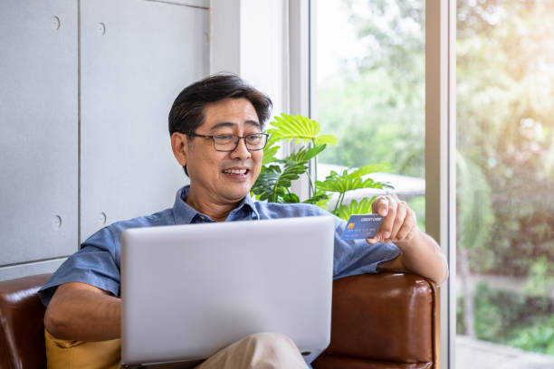 istock Senior Asian man sitting on sofa using laptop computer holding credit card for online shopping and payment 1393605359