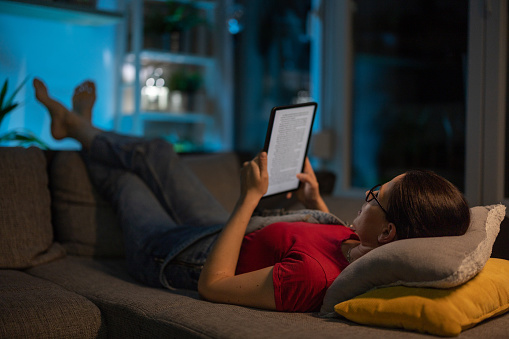 Woman lying on sofa in living room at home, and reading a book on digital tablet in peace.