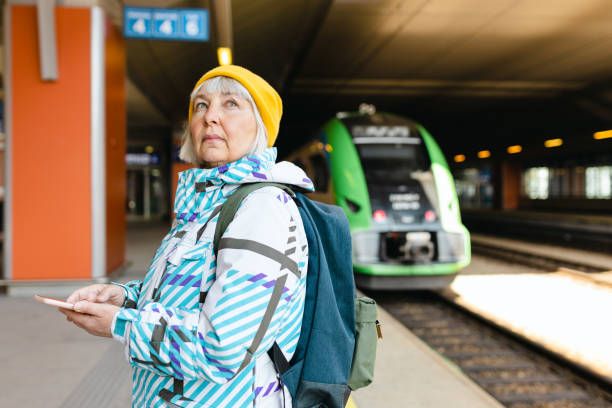 istock Old happy woman with backpack in yellow hat waiting train on station platform and using smart phone on urban background. Railroad transport concept 1393615757