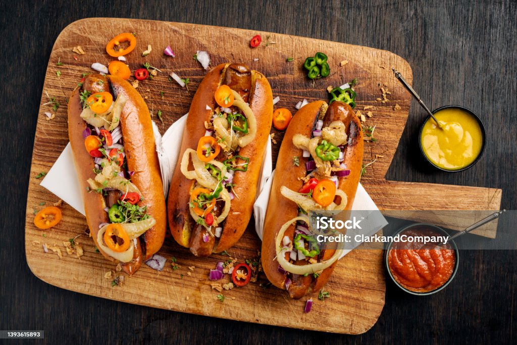 Artisan Hot Dogs in Brioche Rolls with pickles, onion and relishes. Overhead view of three artisan hot-dogs in brioche rolls with pickles and relishes. Colour, horizontal format with  some copy space. Hot Dog Stock Photo