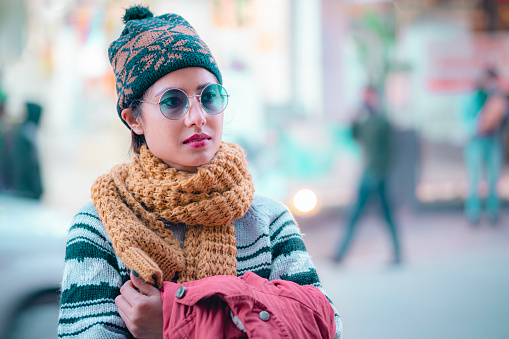 In this winter night image with copy space, an Asian/Indian confident young woman looks away and walks on a brightly lit city street. She wears a warm cap, muffler, sweater, and sunglasses.