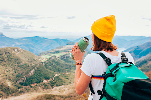 Back view of drinking hiking woman with bottle of water, yellow beanie and backpack. Outdoor sports, adventure, lifestyle, travel, vacations, holiday concept.