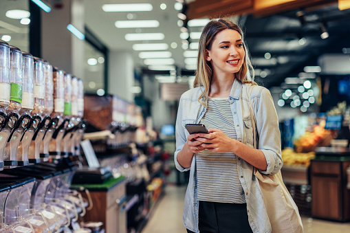 Young woman with shopping bag looking for price of groceries on cellphone