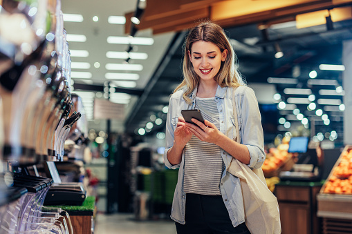 Young female consumer with shopping bag standing by shelf with groceries and using phone, checking shopping list, while choosing goods in supermarket