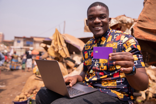 African man shows his credit card while using his laptop, he is at the market. Credit card payments in Africa concept